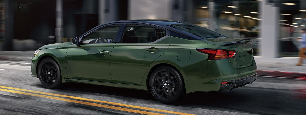 2026 Nissan Altima 3/4 rear side view of  Altima driving on a city road with modern building in background.