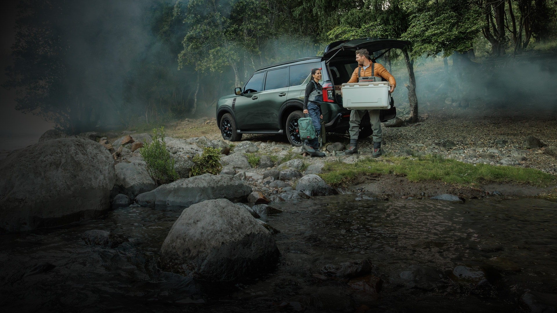 2026 Nissan Armada parked by a river with the cargo space open as two people unload a cooler and backpacks from it